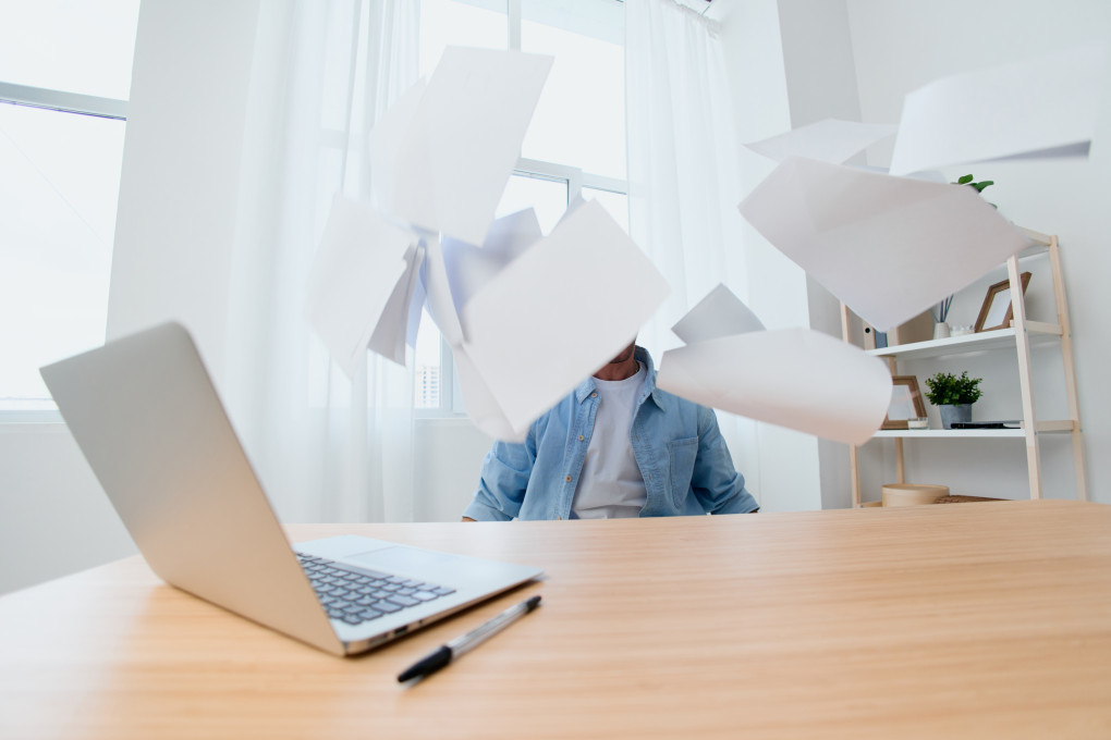 Eine Person sitzt am Schreibtisch, überall fliegen Blätter. Auf dem Schreibtisch steht ein Laptop.
